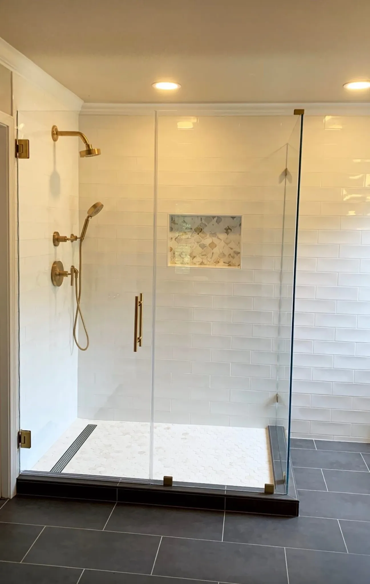 Custom walk-in shower with frameless glass, brass fixtures, linear drain, and recessed mosaic niche